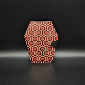 May include: A wallet with a geometric pattern in red and black. The wallet has a black clasp and a unique hexagonal shape. The design is a repeating pattern of interlocking hexagons. The wallet is made of a textured material.