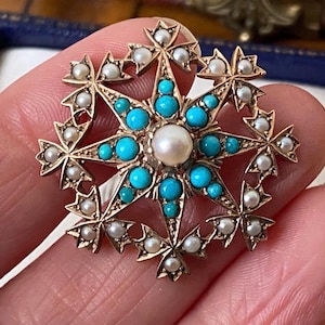 Puede incluir: Un broche en forma de estrella hecho de metal dorado, con una perla blanca central rodeada de cabujones de color turquesa brillante. Las puntas exteriores de la estrella están adornadas con pequeñas perlas blancas, creando un diseño floral simétrico.