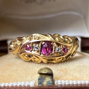 May include: A gold ring featuring a central oval-cut pink gemstone flanked by two small clear stones and two additional pink stones. The band displays an intricate, raised scrollwork pattern across the surface, creating a textured appearance.
