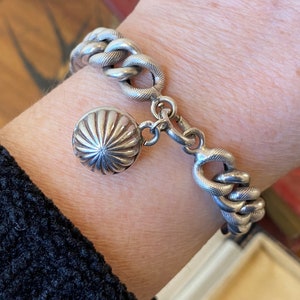 May include: A silver bracelet with a textured chain link design. The bracelet features a decorative round charm with a ribbed pattern. The clasp is a hook and eye style. The bracelet is worn on a wrist.