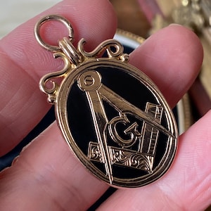 Puede incluir: Colgante dorado con un centro negro ovalado con símbolos masónicos. El colgante tiene un lazo decorativo y detalles ornamentados en los bordes. Los símbolos incluyen una brújula, una escuadra y la letra "G".