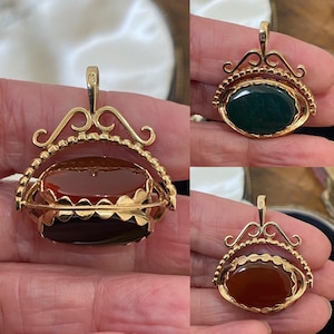 May include: A gold swivel pendant featuring an oval-shaped stone that rotates to reveal two different sides. One side displays a deep red carnelian, while the other side shows a dark green bloodstone. The metal frame has decorative scrollwork.