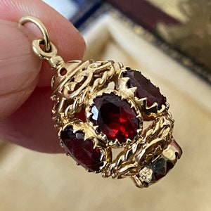 Könnte beinhalten: Goldener Anhänger mit mehreren ovalen, tiefroten Edelsteinen. Der Anhänger hat ein verziertes Filigran-Design und eine kleine Schlaufe oben für eine Kette. Die Edelsteine sind in einen goldenen Rahmen gefasst.