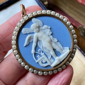 Può includere: Ciondolo rotondo con cammeo azzurro raffigurante una figura classica alata con una lira. Il cammeo è incastonato in una cornice dorata e circondato da piccole perle. Il ciondolo ha un anello dorato nella parte superiore.