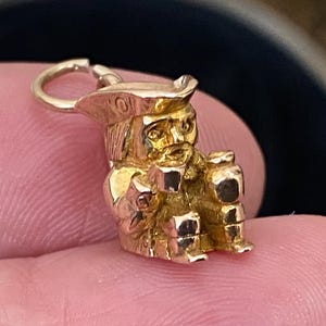 May include: A gold charm shaped like a seated figure wearing a tricorn hat. The figure has a detailed face with a beard and is holding a small object in its hands. A small circular jump ring is attached to the top of the hat for hanging.