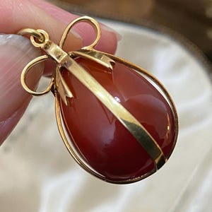 Puede incluir: Colgante dorado con una piedra grande, ovalada, de color marrón rojizo. La piedra está encerrada en una jaula dorada con una parte superior en forma de lazo. El colgante está sujeto por los dedos de una persona.