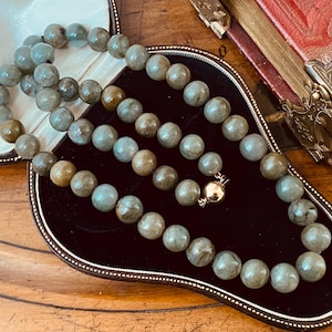 May include: A long necklace made of green and brown gemstone beads. The necklace is in a black velvet box with a gold clasp.