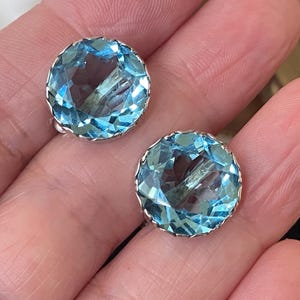 Op de afbeelding: Een paar ronde, lichtblauwe edelsteen oorbellen met een facetontwerp. De oorbellen zijn gezet in een zilverkleurig metaal. De edelstenen hebben een briljante, sprankelende uitstraling. De oorbellen worden op een hand getoond.