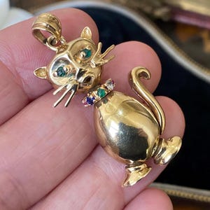 May include: A gold cat pendant featuring green gemstone eyes and a collar set with alternating small blue, green, and clear gemstones. The cat has a polished finish, pointed ears, whiskers, and a curved tail, with a bail at the top for hanging.