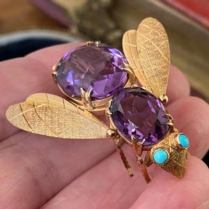 May include: A gold-toned brooch in the shape of a stylized insect. The brooch features two large, oval, purple gemstones for the body and head, with turquoise accents for the eyes. The wings are textured gold.
