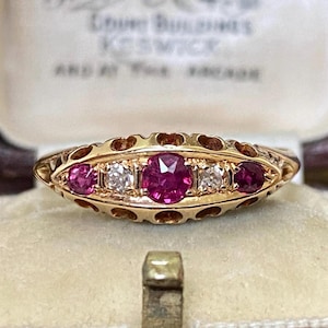 Może przedstawiać: Złoty pierścionek z czerwonymi i przezroczystymi kamieniami szlachetnymi. Pierścionek osadzony jest w ozdobnej oprawie z szeregiem małych, wyciętych kształtów. Pierścionek prezentowany jest w zabytkowym pudełku na biżuterię.