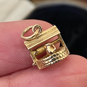 May include: A gold-colored charm in the shape of a well. The charm features a detailed brick-like design around the base and a small handle. The well's structure includes a small, round opening and a detailed winding mechanism.