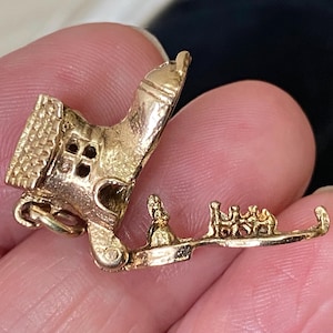 May include: A gold charm in the shape of a shoe that opens to reveal a miniature interior scene. The exterior features a textured roof and window details, while the interior base displays small figures of a person and a group of children.