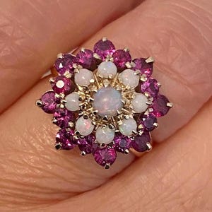 May include: A cluster ring featuring a central round opal surrounded by a halo of eight smaller round opals, encircled by an outer layer of twelve faceted deep pink gemstones set in a gold-toned metal frame with a prong setting design.
