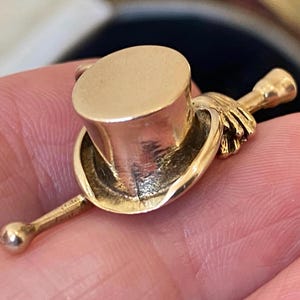 Puede incluir: Un dije dorado con forma de sombrero de copa y bastón. El dije tiene un diseño detallado, con el sombrero y el bastón conectados por una mano. El dije probablemente está hecho de oro o un metal dorado.