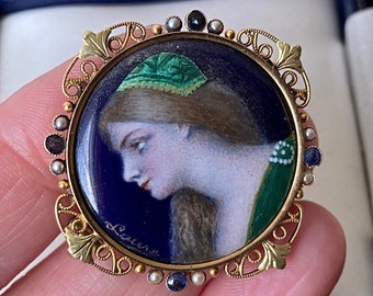 Könnte beinhalten: Eine goldfarbene Brosche mit blauem Emaille-Hintergrund und einem gemalten Porträt einer Frau mit grünem Kopftuch. Die Brosche ist mit einem filigranen Design und Perlen verziert.