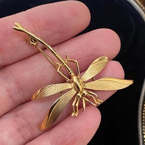 May include: A gold-toned dragonfly brooch featuring a textured finish on the wings and a smooth, polished body. The insect-shaped pin has a long, slender tail and delicate legs, with a functional metal clasp mechanism visible on the reverse side.
