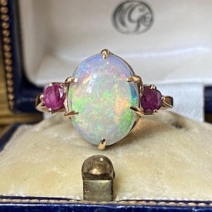 May include: A gold ring featuring a large oval cabochon opal with iridescent flashes of green and orange, flanked by two round faceted deep pink rubies. The stones are set in a gold band with four prongs securing the central opal gemstone.