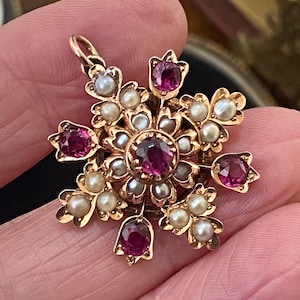 Puede incluir: Un colgante de oro con una piedra preciosa roja redonda central rodeada por un grupo de pequeñas perlas blancas. El diseño incluye piedras rojas adicionales y detalles de perlas dispuestos en un patrón floral o de estrella con una anilla.