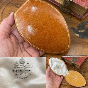 Op de afbeelding: Een ovaalvormig beige lederen juwelendoosje met goudkleurige reliëfrand. De binnenzijde is bekleed met wit satijn en voorzien van het gedrukte logo van Alfred Speed, Goldsmith & Diamond Merchant, 84 High St, Kings Lynn, met een kroonicoon.