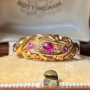Pode incluir: Um anel de ouro com uma pedra preciosa rosa redonda central, ladeada por duas pedras claras e duas pedras rosas menores. A banda tem um design intrincado de volutas vazadas. O anel é apresentado numa caixa com o texto NOTTINGHAM visível.