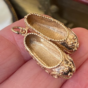 Może przedstawiać: Złoty metalowy charms w kształcie pary miniaturowych butów. Buty mają teksturowane wykończenie, sznurowane zdobienie wzdłuż otworu oraz małe ozdobne ćwieki na zaokrąglonych noskach. Z boku przymocowane jest małe kółeczko do zawieszenia.