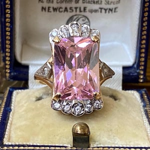 Könnte beinhalten: Ein Goldring mit einem großen, rechteckigen, facettierten rosa Edelstein in der Mitte. Der Stein ist von einem Kranz aus kleinen, klaren, rund geschliffenen Steinen in weißem Metall umgeben, mit weiteren kleinen Steinen an den Seiten.