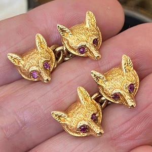 Puede incluir: Un par de gemelos de metal en tono dorado, cada uno con dos motivos esculpidos de cabeza de zorro conectados por una cadena corta. Cada cabeza de zorro tiene una superficie texturizada y una pequeña piedra preciosa redonda de color rosa intenso como ojo.