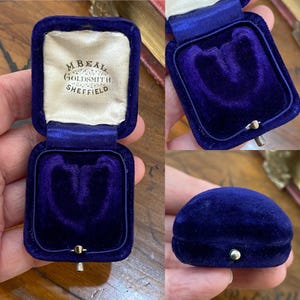 May include: A square, deep blue velvet jewelry box with a hinged lid. The interior features a cream-colored satin lining printed with the text M. BEAL GOLDSMITH SHEFFIELD. The base is lined with dark purple velvet and includes a small metal latch.