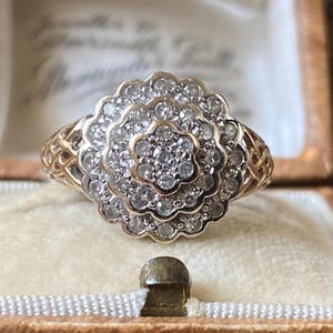 Op de afbeelding: Een gouden ring met een grote, bloemvormige diamantcluster. De ring is gezet in een delicaat, filigraan ontwerp.