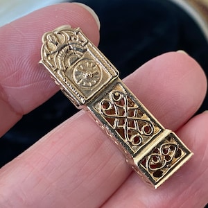 Op de afbeelding: Een goudkleurige, beweegbare bedel in de vorm van een miniatuurklok. De bedel heeft ingewikkelde filigraan en een wijzerplaatontwerp. De bedel wordt in een hand gehouden.