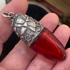 May include: A vintage perfume bottle featuring a silver-tone cap and a vivid red glass body. The cap is adorned with detailed floral and insect designs. The bottle has a loop for a chain, indicating it could be worn as a pendant.