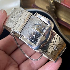 Puede incluir: Un brazalete articulado de metal plateado con un mecanismo de cierre tipo hebilla. La superficie del brazalete está decorada con intrincados patrones grabados e incluye una cadena de seguridad fina y delicada en ambos lados.