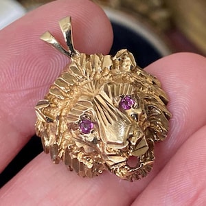 Peut inclure: Pendentif en or en forme de tête de lion avec une surface texturée et deux petites pierres roses rondes pour les yeux. Le pendentif a une boucle en haut pour une chaîne. Le visage du lion est détaillé.