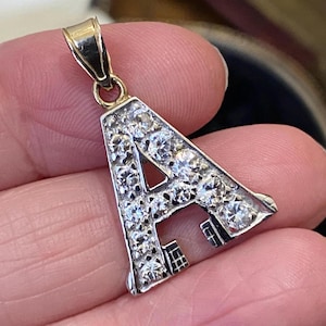 Puede incluir: Un colgante con forma de la letra "A", incrustado con numerosas piedras preciosas pequeñas y brillantes. El colgante tiene un marco plateado y una fianza dorada. Las piedras preciosas claras reflejan la luz, creando un efecto deslumbrante.