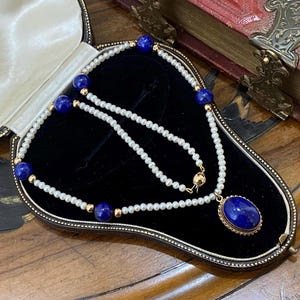 Puede incluir: Un collar vintage con colgante y cadena de perlas, presentado en un joyero de terciopelo negro. El collar presenta un gran colgante ovalado de piedra azul y piedras azules más pequeñas intercaladas con perlas.