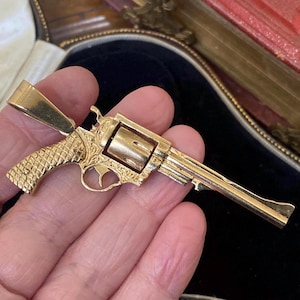 May include: A gold-colored metal pendant shaped like a revolver firearm. The pendant features a textured grip, an engraved frame, a rotating cylinder, and a long barrel. A large, smooth, tapered bail is attached to the top of the handle for hanging.