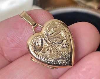 May include: A gold-toned heart-shaped locket pendant featuring an intricate engraved floral pattern on the front surface. The locket includes a small, rectangular bail at the top for attachment to a chain, with a polished finish and visible hinge.