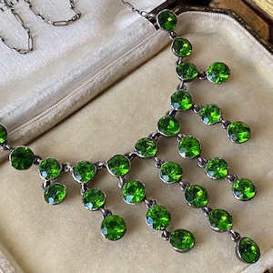 Puede incluir: Un collar de plata vintage con una cascada de piedras preciosas verdes vibrantes. El collar presenta una delicada cadena y múltiples hilos de piedras redondas y facetadas, creando un diseño brillante y elegante. El collar se exhibe en un joyero vintage.