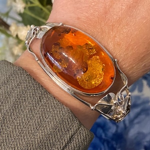 Op de afbeelding: Een zilveren armband met een grote, ovale, doorschijnende barnsteen met zichtbare gouden insluitingen. De metalen band is afgewerkt met een gepolijste glans en decoratieve, organisch gevormde zilveren accenten aan beide zijden.