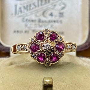 May include: A gold ring with seven deep red gemstones, possibly rubies, encircled by smaller clear stones. The ring has a floral design and is set in a gold band. The ring is displayed in a vintage jewelry box. The ring's design is intricate.