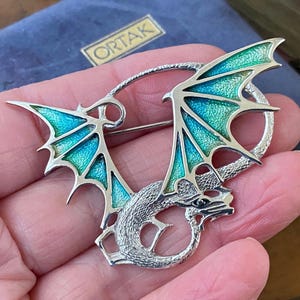 May include: A silver-toned dragon brooch with turquoise and teal enamel wings. The dragon has a detailed, textured body and is set against a blue velvet background. The brooch is a unique piece of jewelry.
