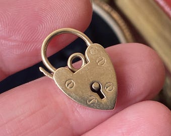 May include: A small, heart-shaped gold padlock with a keyhole and a circular loop. The padlock has a simple design with visible screw heads and a slightly aged appearance, suggesting it is vintage or antique.