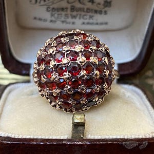 Puede incluir: Un anillo de oro vintage con un grupo de pequeñas piedras preciosas redondas de color rojo oscuro. El engaste del anillo es dorado y las piedras preciosas están muy juntas, formando una forma de cúpula. El anillo se muestra dentro de un joyero marrón.