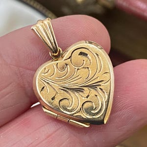 Puede incluir: Un colgante de relicario en forma de corazón de tono dorado con un intrincado patrón de volutas grabado en la superficie frontal. El colgante incluye una pequeña anilla estriada en la parte superior para sujetarlo a una cadena, con un acabado metálico pulido.