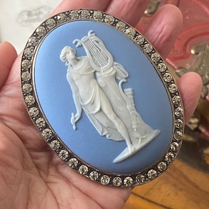 May include: Oval brooch with a light blue cameo featuring a white classical figure with a lyre and bow and arrow. The cameo is surrounded by a border of clear, sparkling rhinestones set in a silver-tone metal frame.