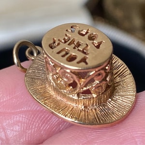 May include: A gold-toned metal charm shaped like a top hat with a textured brim and a filigree band. The flat top of the hat features the embossed text 'YOU THEY ADS'. A small circular jump ring is attached to the side for hanging purposes.