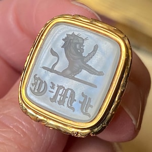 May include: A gold signet ring featuring a rectangular, translucent white stone inlay. The stone is engraved with a dark, detailed image of a lion standing on its hind legs and the gothic-style letters D, M, and L positioned below the lion.