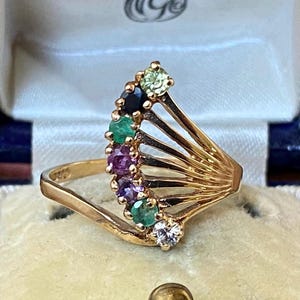 Puede incluir: Un anillo dorado con un diseño en forma de abanico, adornado con una fila de piedras preciosas de colores. Las piedras preciosas incluyen piedras verdes, moradas y blancas. El anillo está sobre un fondo crema.