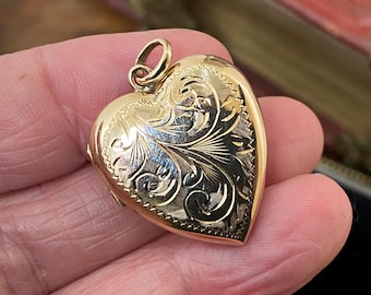 May include: A gold-toned heart-shaped locket pendant featuring an intricate engraved floral pattern on the front surface. The locket has a polished finish with detailed line work and includes a small circular bail at the top for attachment to a chain.